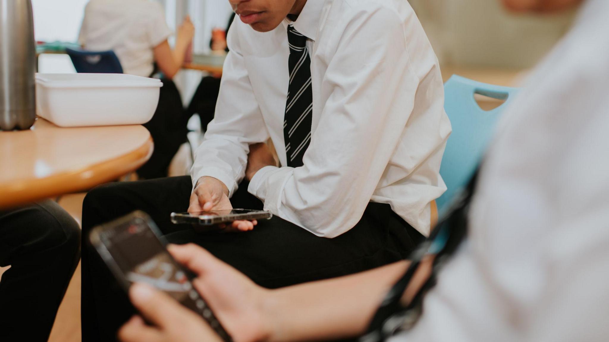 Two students in school uniform sit indoors during a break, looking down at smartphones held in their hands.