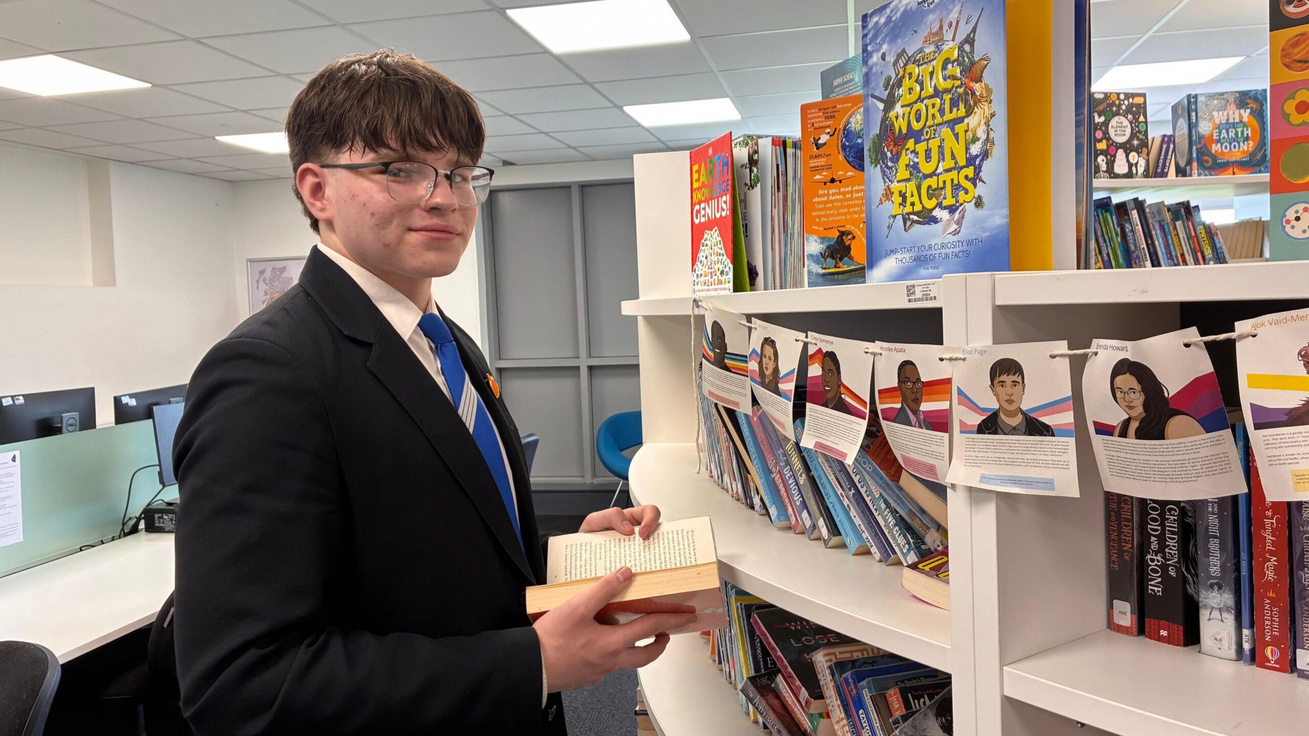 A white teenage boy with brown hair and wearing glasses and school uniform is holding a book in a school library on the left of the picture. He is standing in front of a curved bookcase with computer work stations behind him.