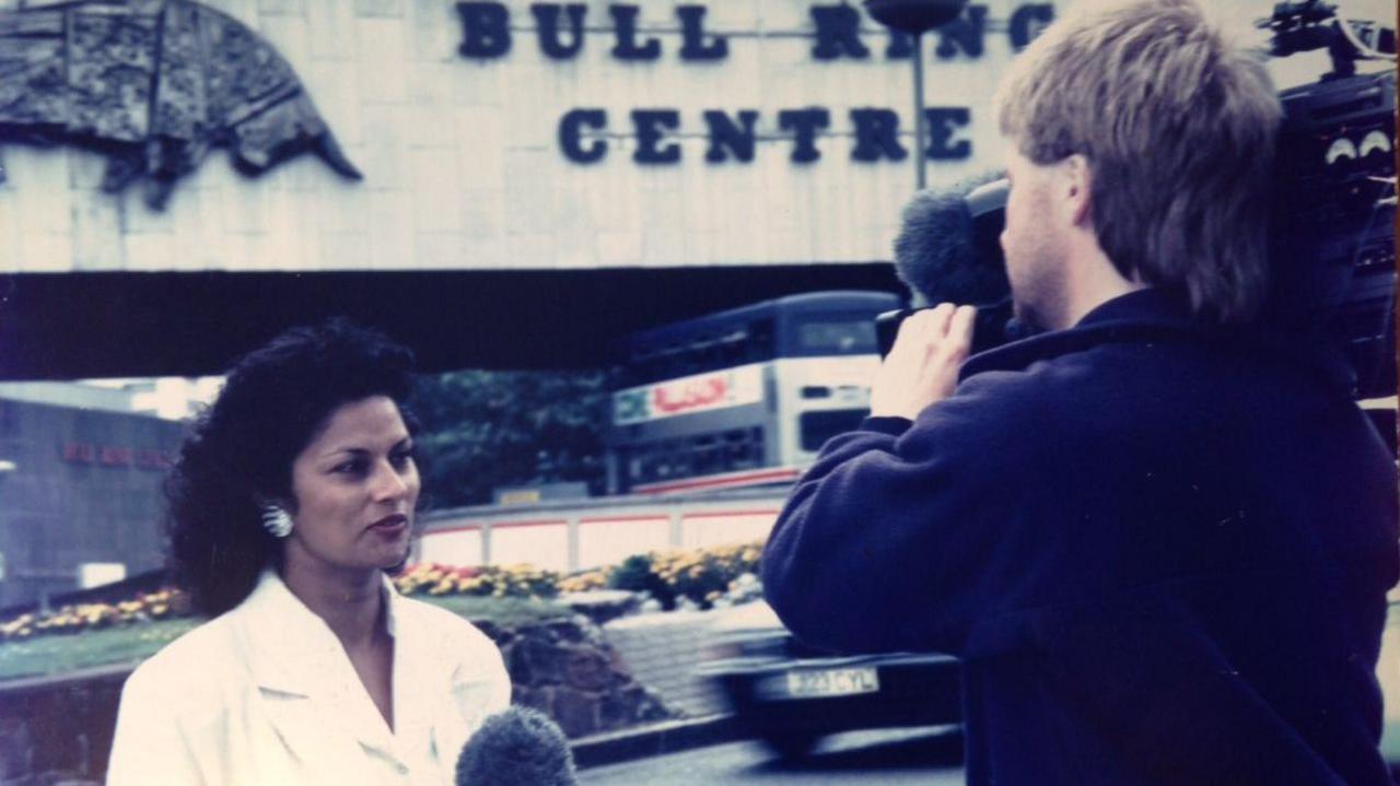 A female news reporter in a white outfit is being filmed by a male camera operator outside a building. Behind them is a sign that reads “Bull Ring Centre” with a metal bull sculpture above it, and traffic and flowerbeds in the background.