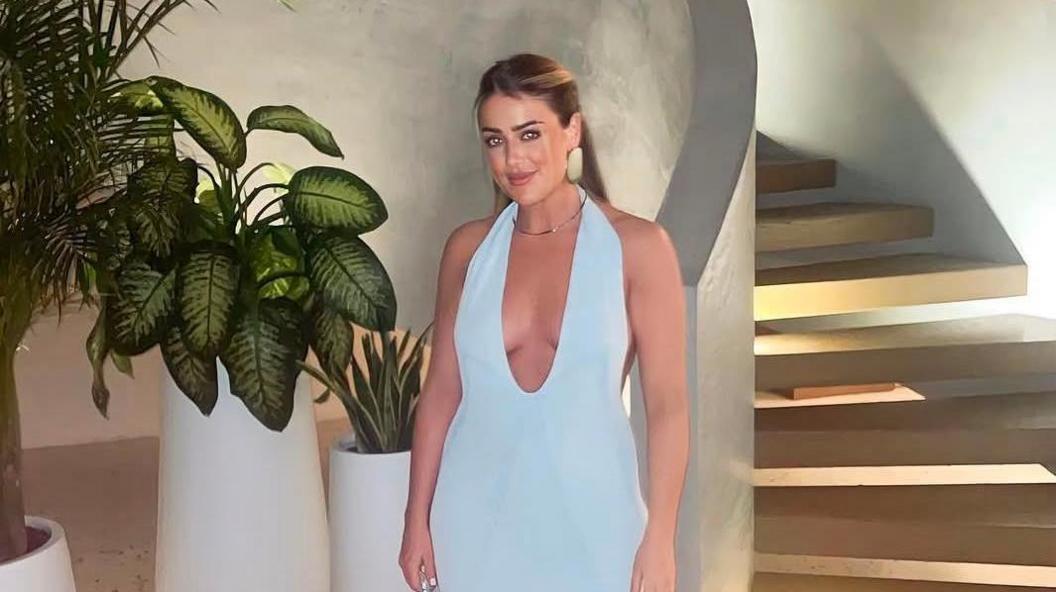 Olivia, a blonde haired woman wearing a light blue, v plunge neckline maxi dress stood in front of stone stairwell.