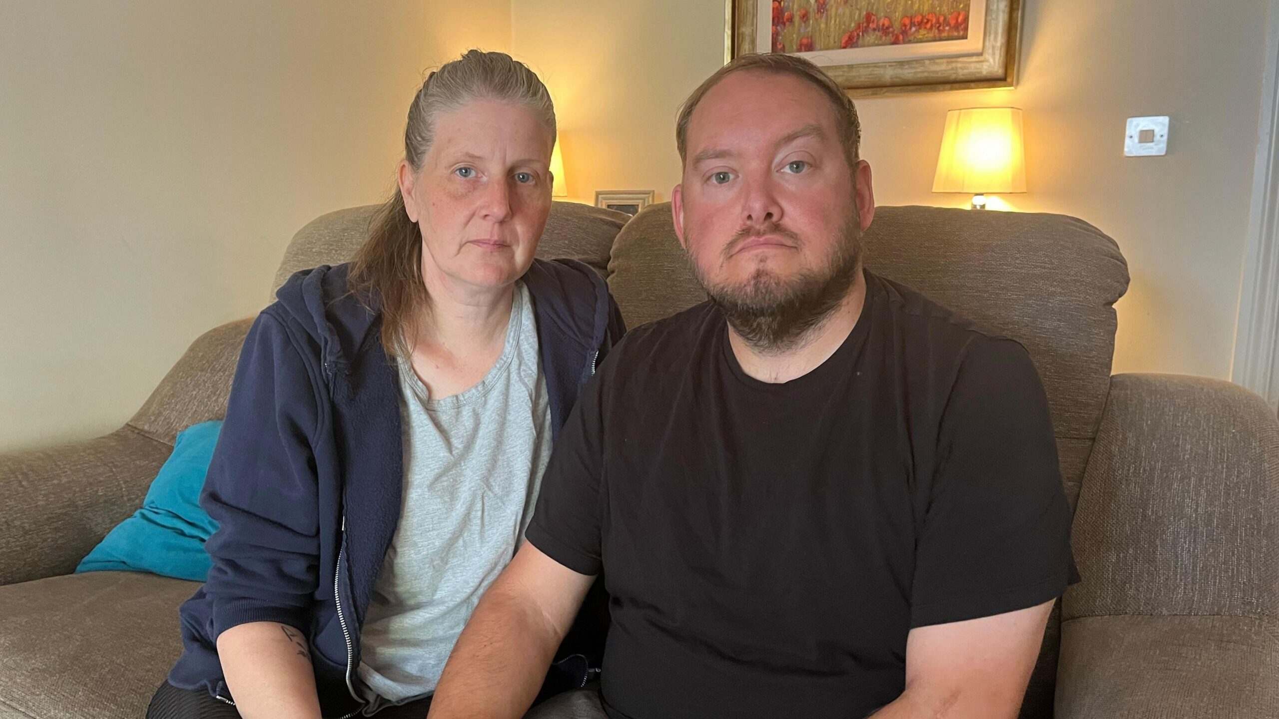 Gary and Sarah are sat on a sofa. Gary is wearing a black t-shirt and Sarah is wearing a grey t-shirt and navy jacket.
