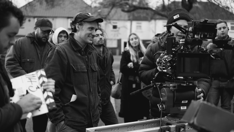 A black and white photo of a man wearing a denim jacket, baseball hat and headphones. Beside him is a woman in a beanie operating a large camera. Behind them, about half a dozen other people look on. There are also houses in the background.