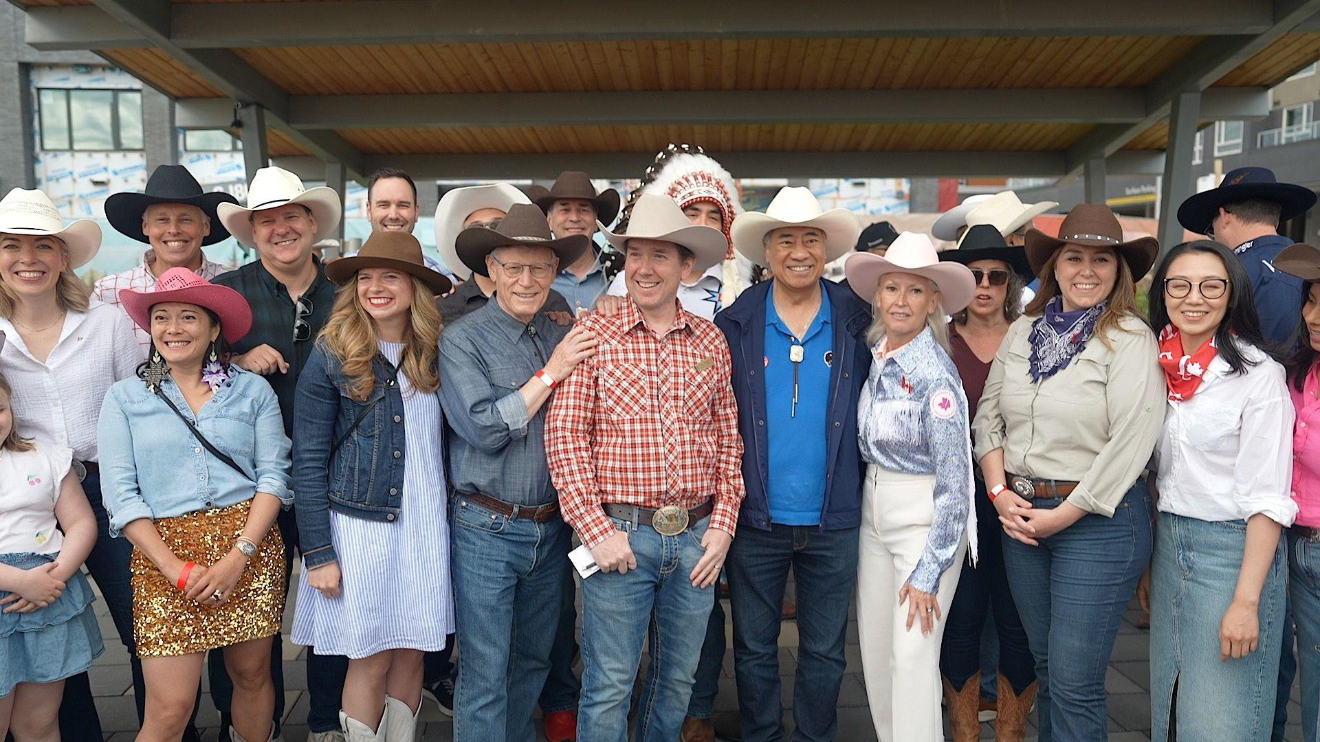 Corey Hogan, centre, in a red and white plaid shirt, a white cowboy hat, and jeans, smiles and poses with dozens of people, also in western regalia.