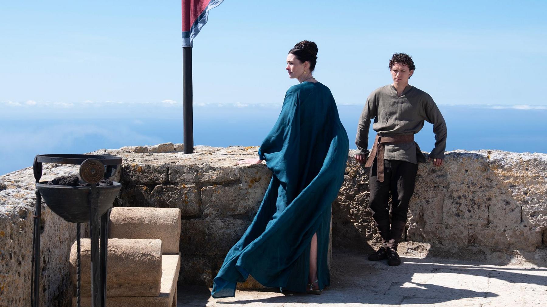 Anne Hathaway as Penelope (Odysseus's wife) in centre of image in blue gown looking to the side and Tom Holland as their son Telemachus leaning on wall in khaki top and dark trousers. Blue skies behind