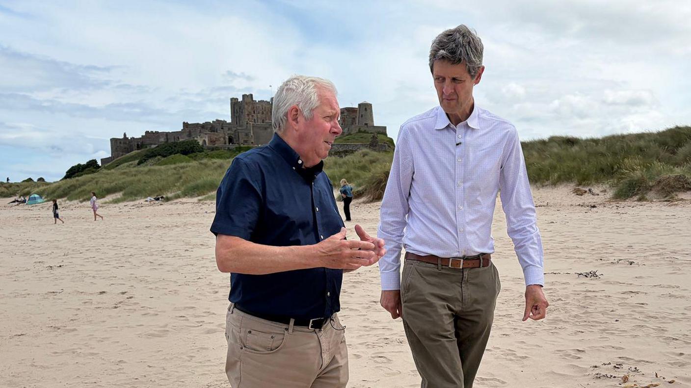 Two men in their 60's are walking down a wide sandy beach. In the background is Bamburgh castle and there are a few clouds in the sky. The sea is calm and the men are clearly engaged in conversation. Both are wearing trousers and relaxed shirts.