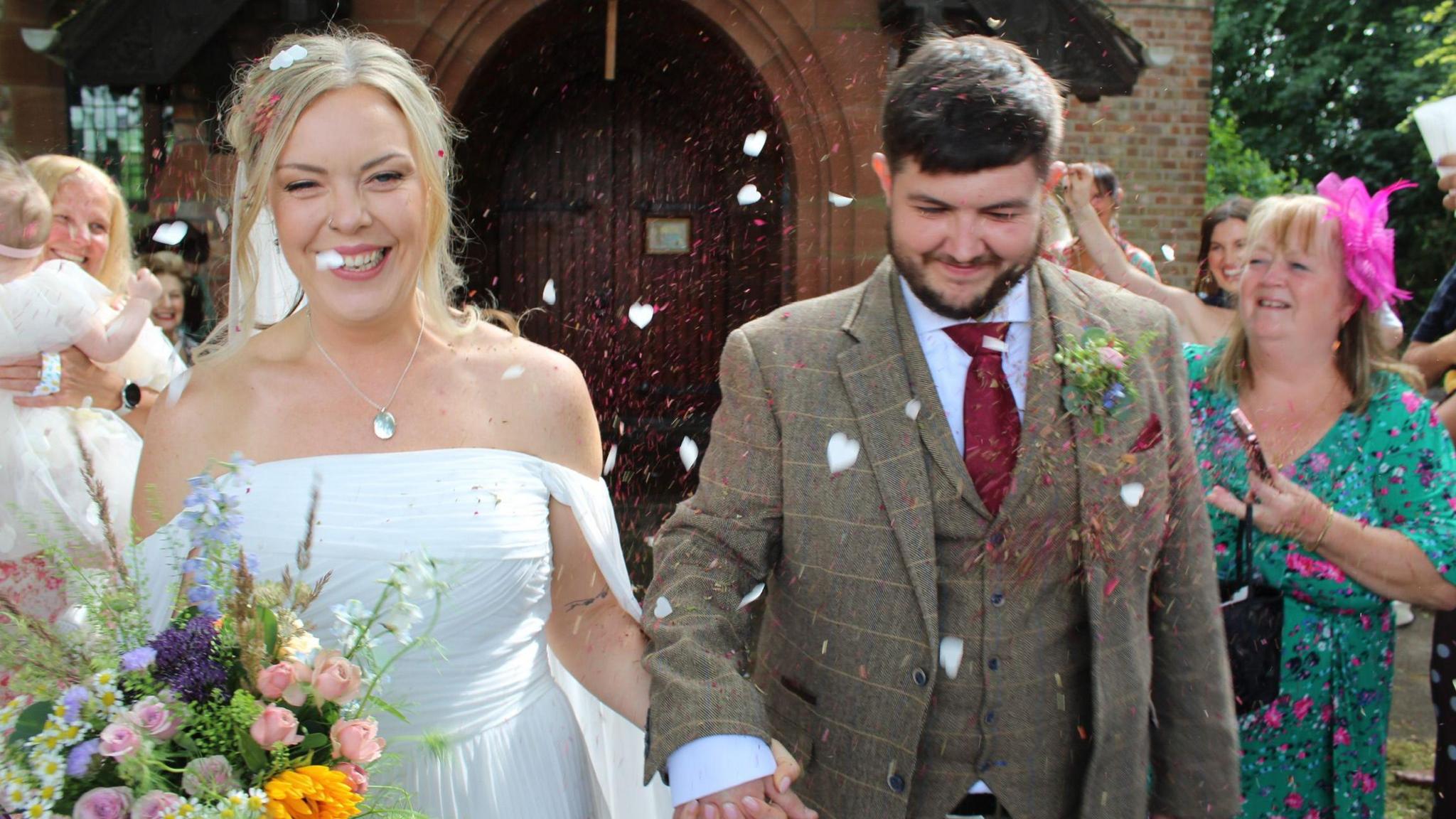 A married couple are pictured smiling, leaving a church, and friends and family are throwing confetti over them. The bride, on the left, is wearing a white dress and carrying flowers. The groom is wearing a moss green checked suit with waistcoat.
