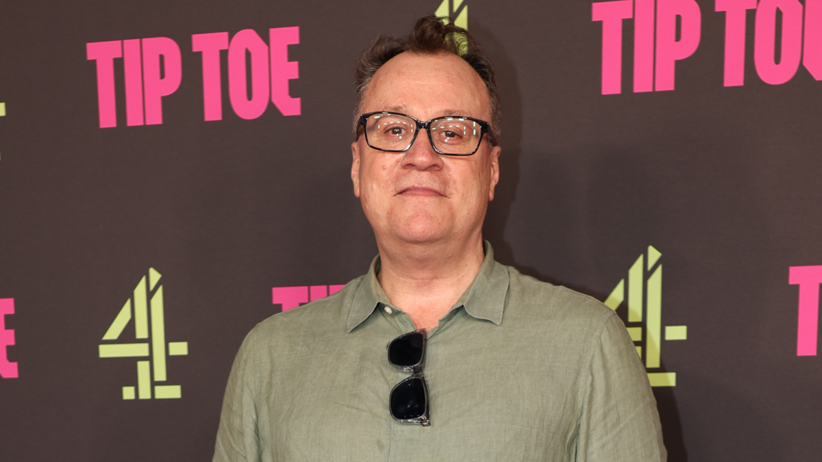 Russell T Davies stands on a red carpet. He wears glasses and short dark hair, and wears a khaki green shirt with sunglasses tucked in the collar.
