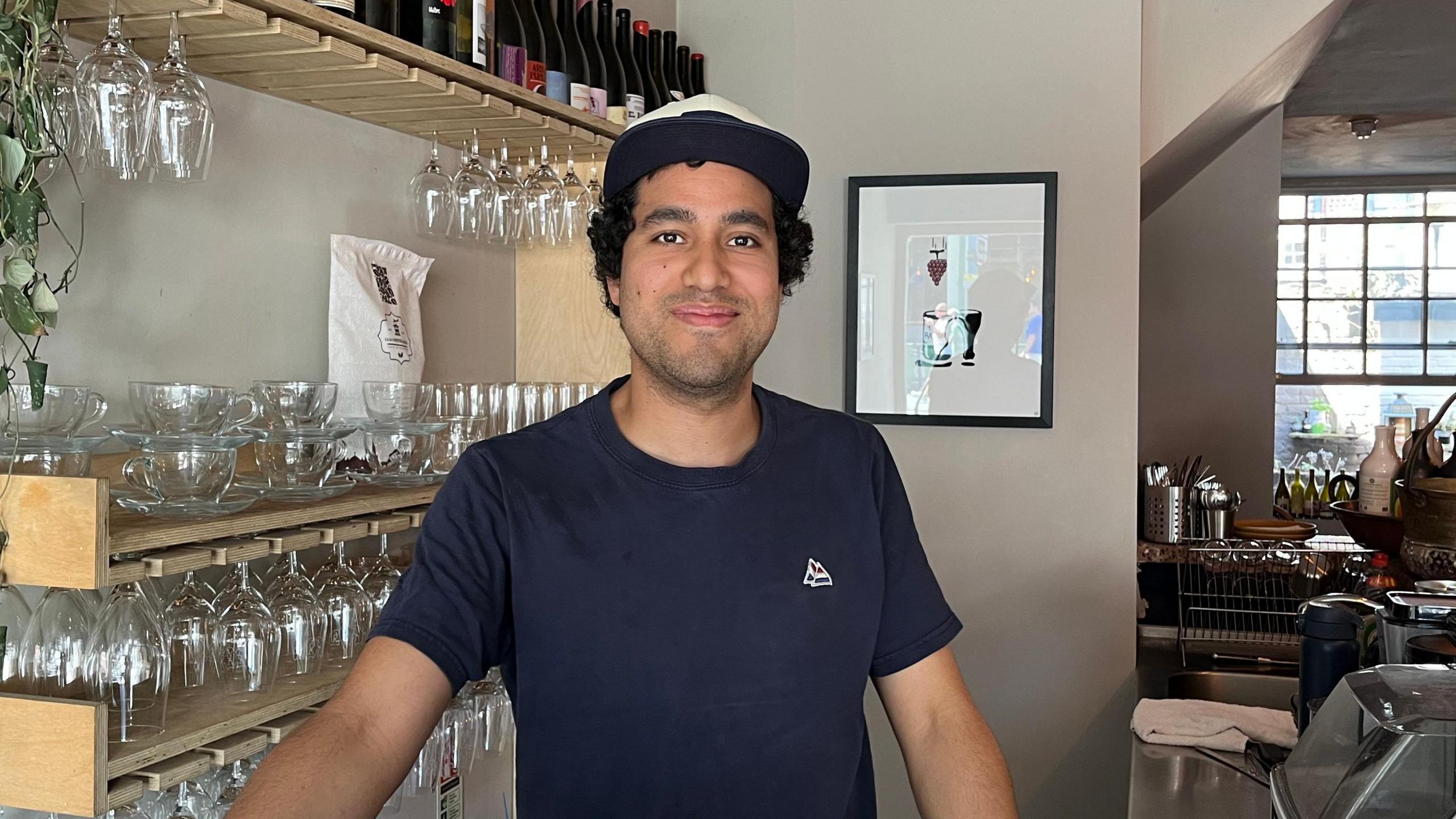 A man stands behind a café counter, with shelves of glassware and wine bottles, hanging lights and food displays visible around the venue.