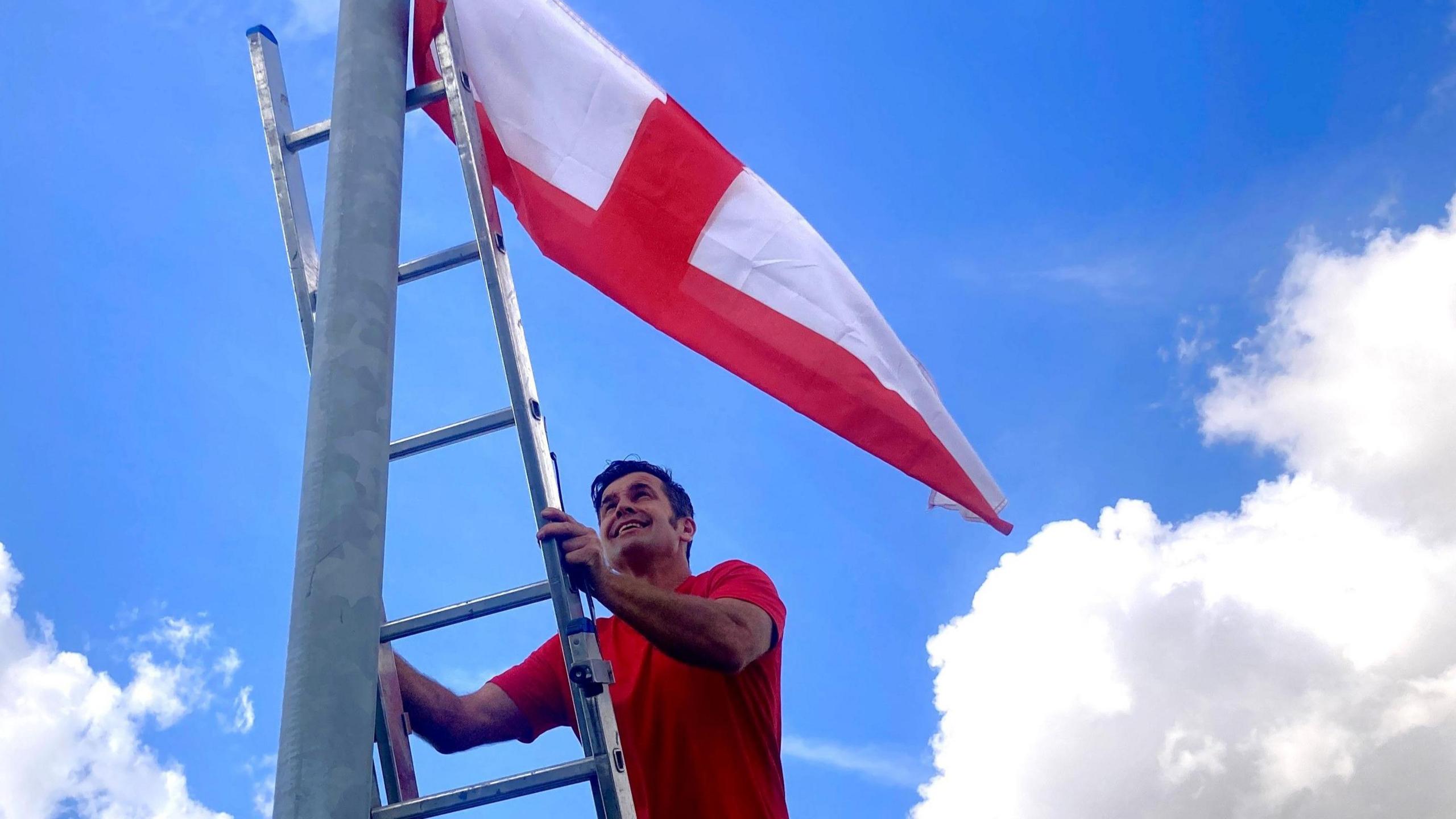 A man wearing a red T-shirt climbing a ladder to fix a St George's flag to a lamp-post.
