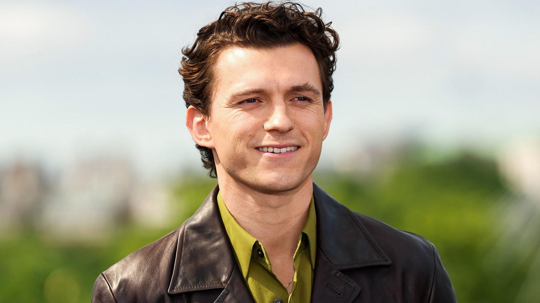 Tom Holland smiling in an olive green shirt and brown leather jacket standing in front of  some green foliage in the distance and a blue sky in a photoshoot for the film The Odyssey in London