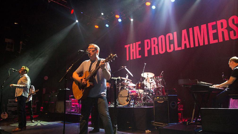 The Proclaimers performing on stage. The two brothers are singing into mics and there are people playing the drums and keyboards behind them. There is a large projection saying The Proclaimers in red