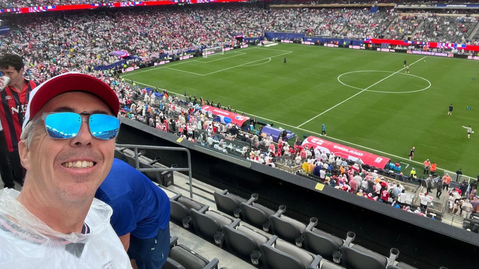 Kevin Craig, wearing a cap and sunglasses, smiles in a selfie with a football pitch in the background.
