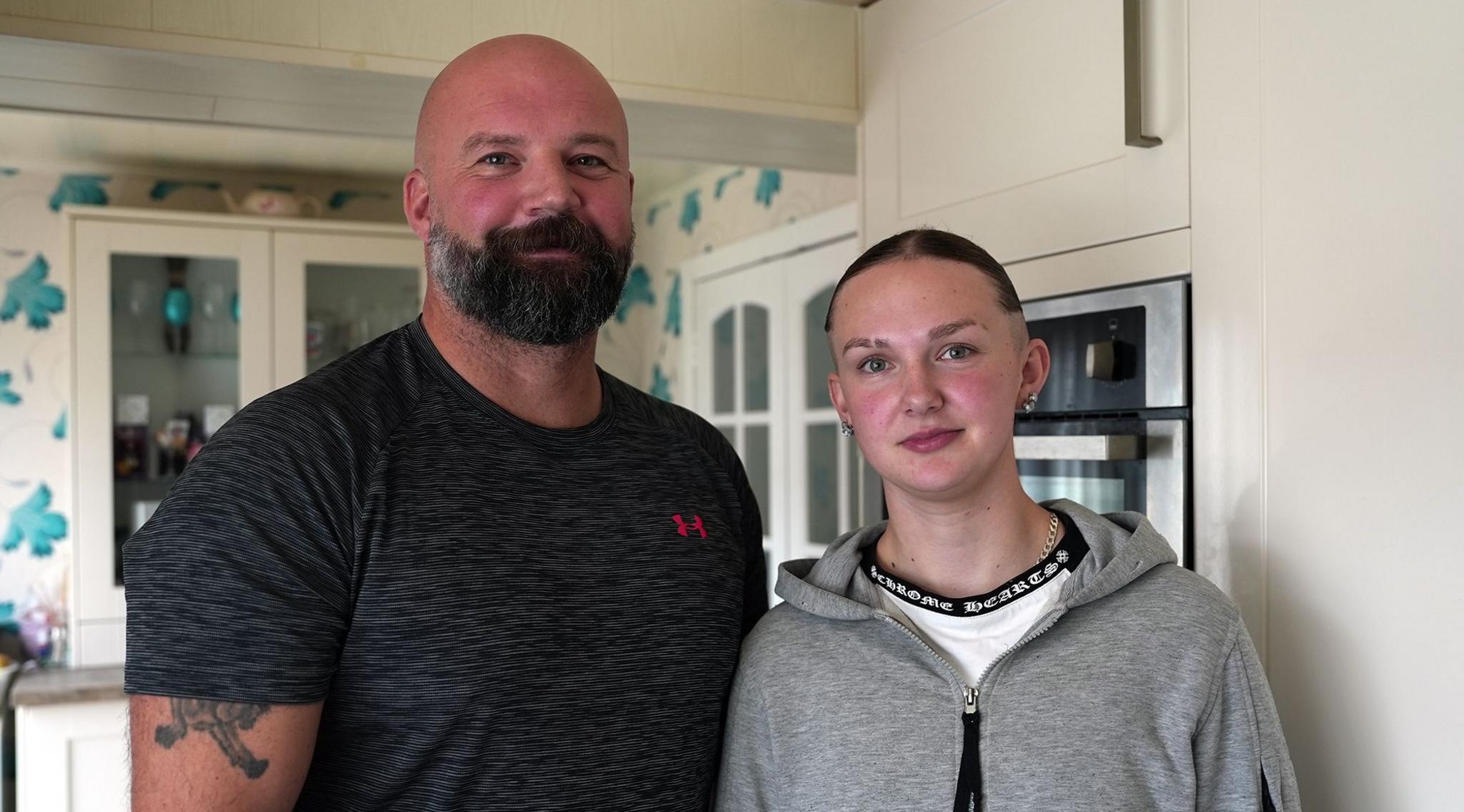 A well built man wearing a dark T shirt stands beside a young woman who is wearing a grey hoodie