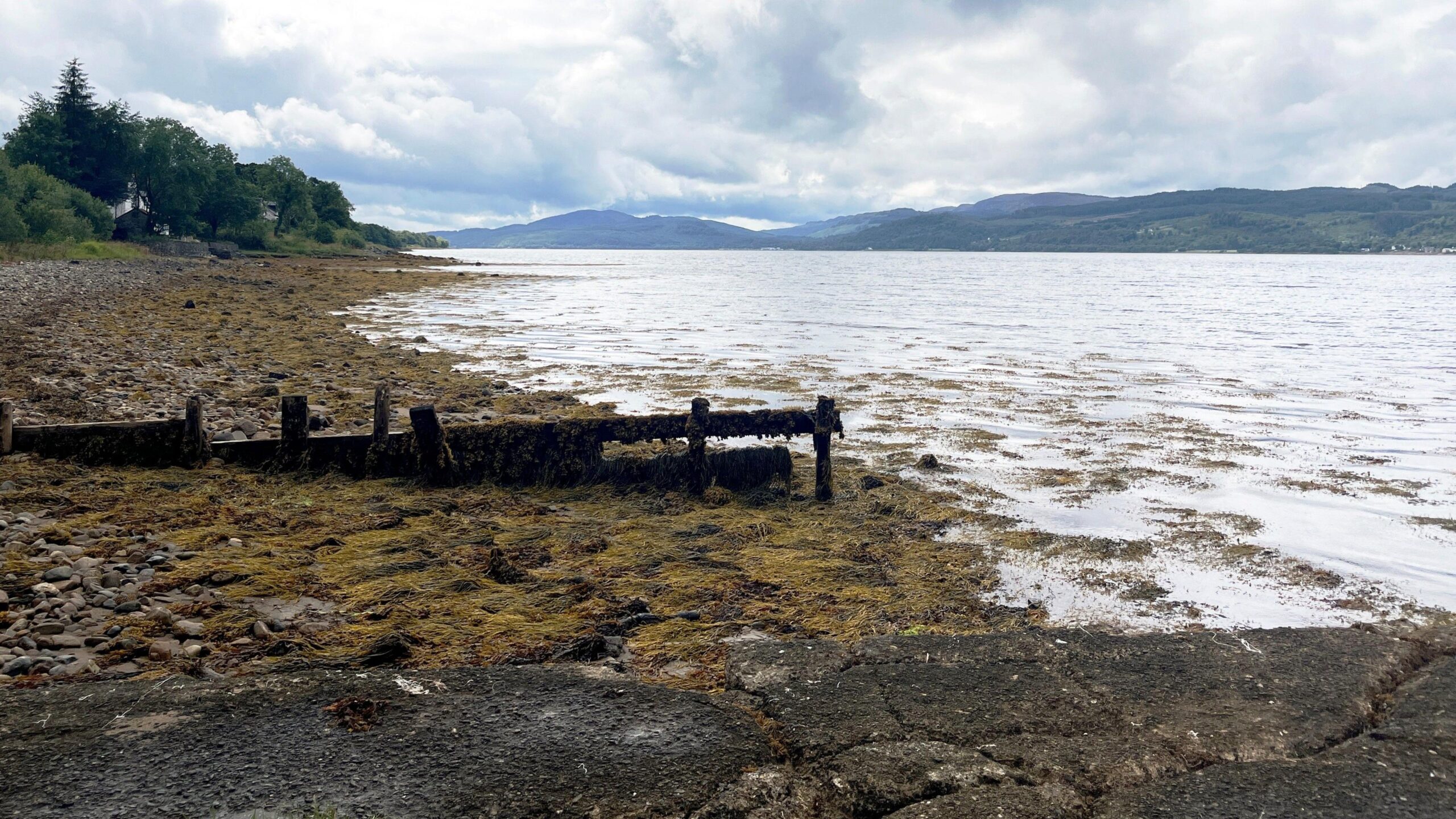 A muddy area leads down a gentle slop into a loch with mountains in the background on a cloudy day