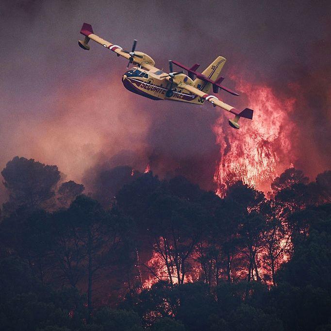 A small yellow airplane flies over red flames rising in smoke above dark trees.
