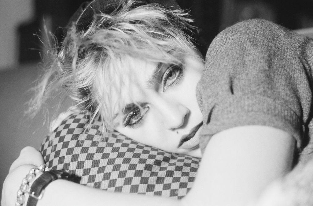 A black and white promotional image of Madonna in 1982 shows the singer lying on a chequered pillow and gazing into the camera, while her artfully messy hair shows dark roots under bleached blonde tips