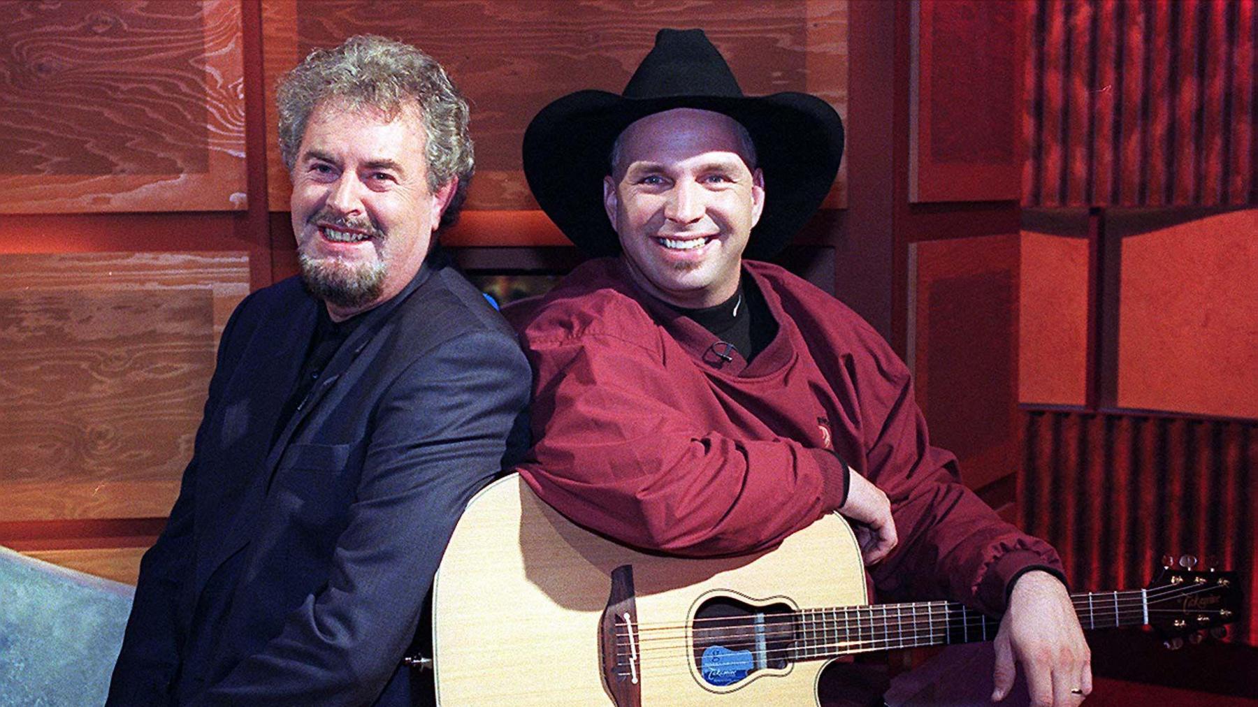 Two men sitting side by side. One has short grey hair and beard, wearing a black blazer. The other is wearing a cowboy hat and has a red shirt on and a guitar on his lap.