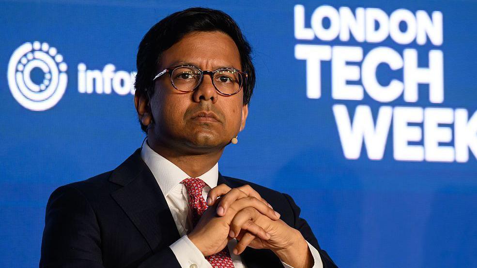 Kanishka Narayan looks pensive with his hands folded together and a microphone positioned by his chin as he sits on stage at London Tech Week. The conference's logo is displayed on a backdrop behind him.