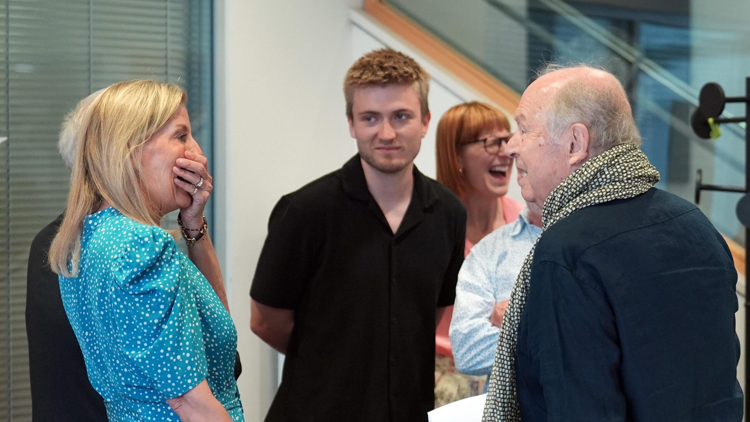 A group of four people are gathered in an indoor setting, The woman on the left, Sophie, wearing a blue polka dot dress, is holding her hand over her mouth while laughing. A man in a black shirt stands behind her, to the right of him, a woman in a pink top laughs heartily. In the foreground on the right, a man with a scarf is speaking to the woman in the blue dress.