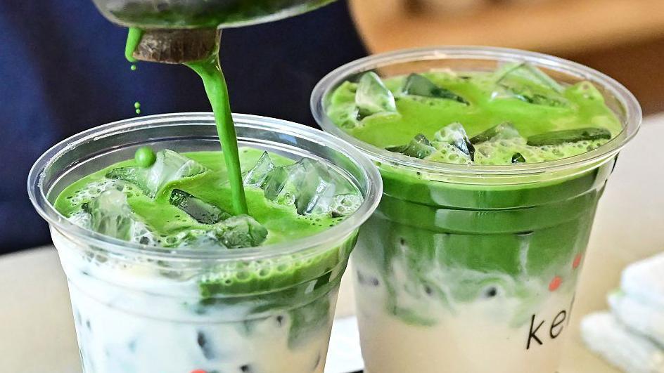 The photo shows two cups of matcha latte being prepared, with the bright green tea being poured into one of the plastic cups filled with milk.