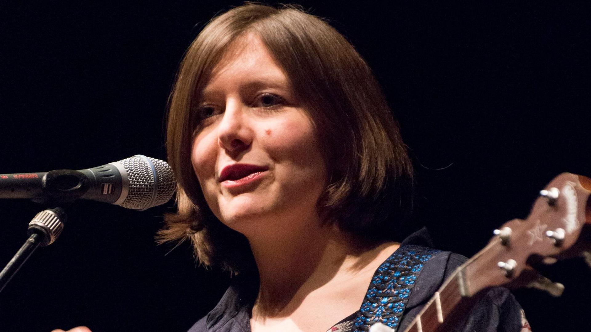 Emily Portman holding a guitar and speaking into a microphone on stage