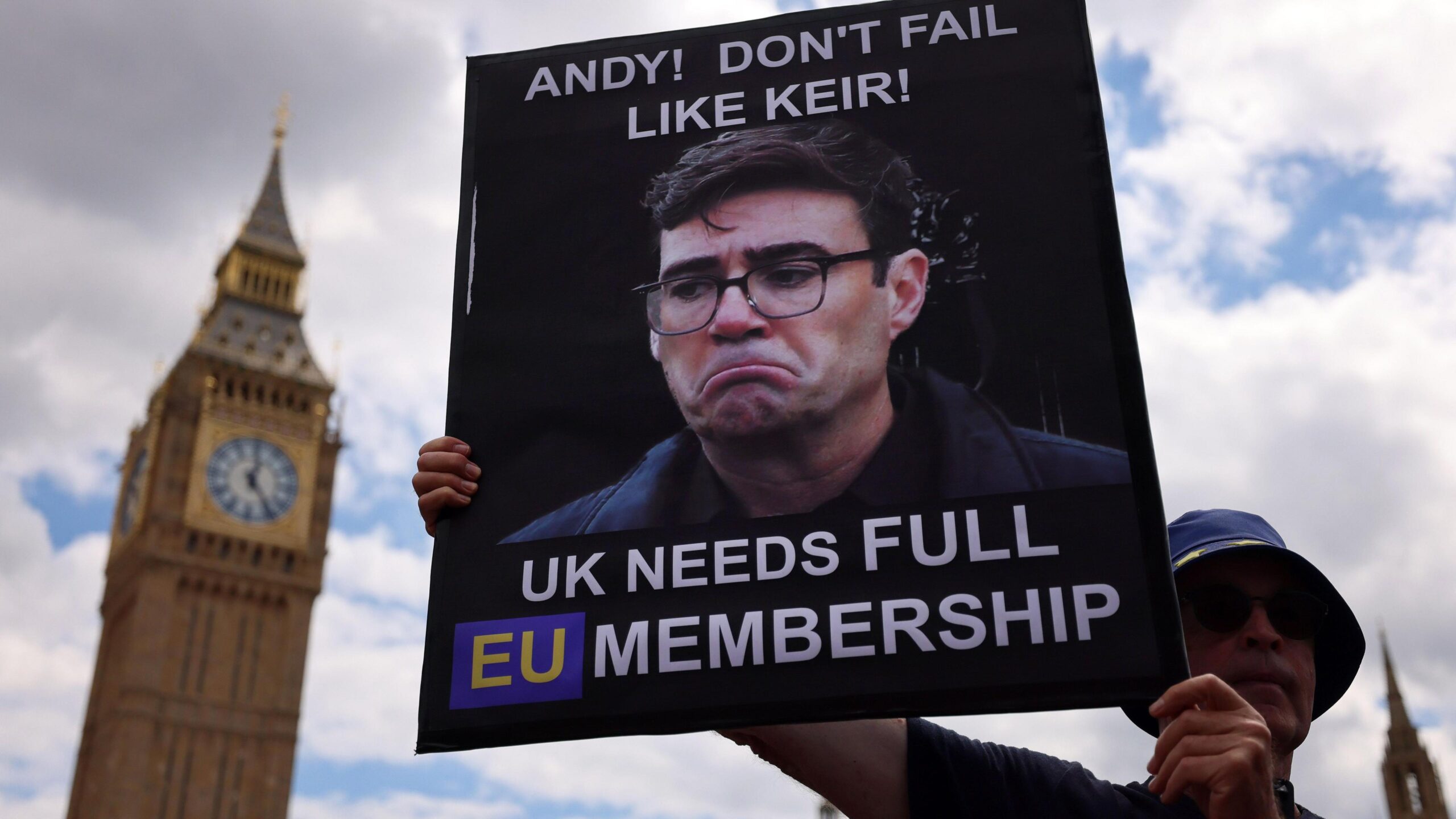 A pro-EU campaigner stands outside Parliament holding up a sign picturing Andy Burnham with a down-turned mouth. The sign says: "Andy! Don't fail like Keir! UK needs full EU membership"