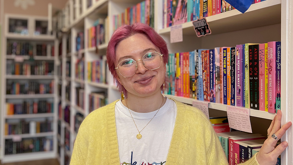 Caden Armstrong is a white women in her 20s. She has pink dyed short to neck-length hair, wears large wire-framed glasses with long dangly gold earrings ending in discs and a small gold medallion necklace. She wears a white t-shire with the top of some writing visible and a yellow cardigan over it. Her left hand rests against a bookshelf and  has two silver rings visible. Behind her are rows of white bookshelves with books filling them, slightly out of focus as the background recedes.