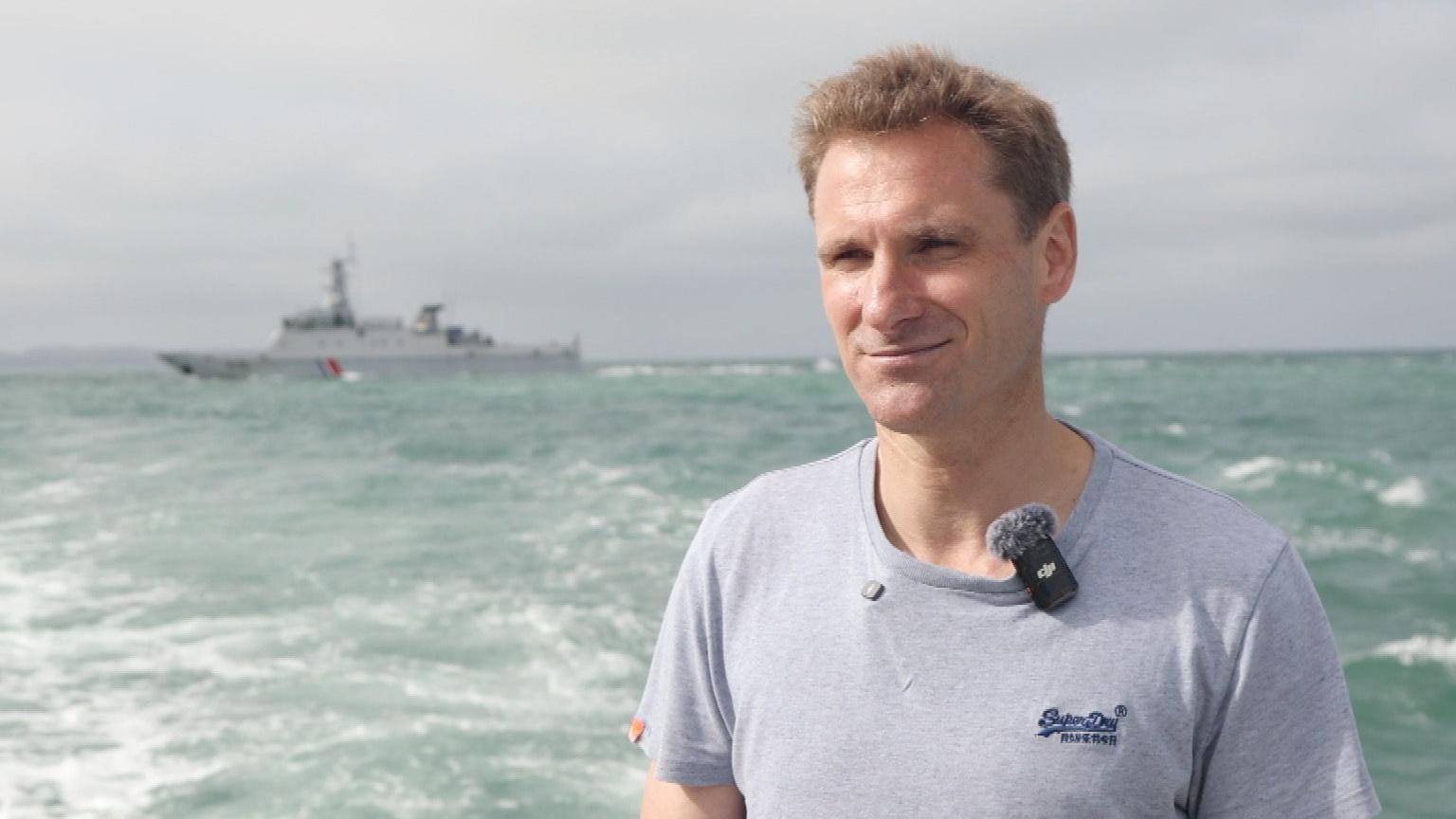 MP Chris Philip, in a grey t-shirt, stands on a boat in the sea. A large, grey ship is visible in the background.