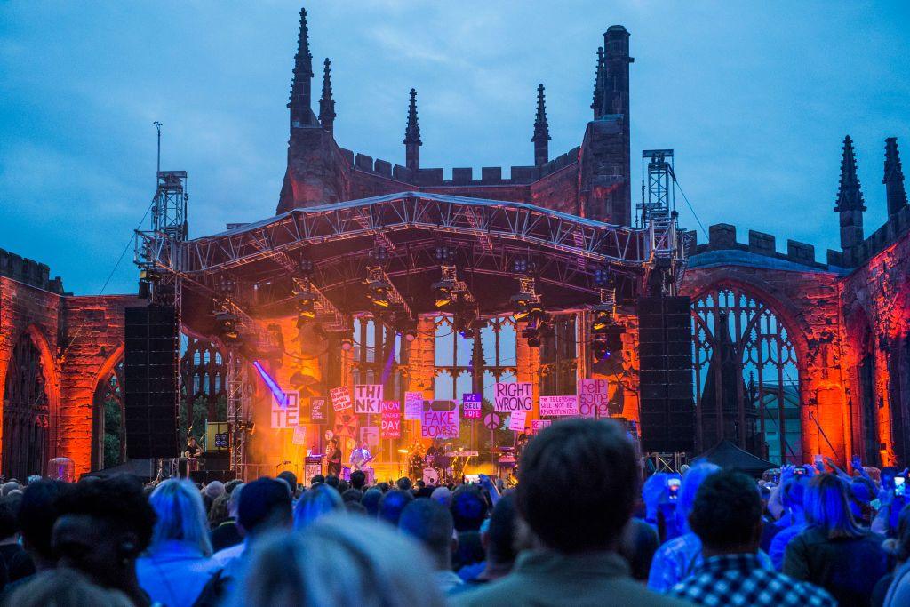 The ruins of Coventry Cathedral can be seen behind a stage which it lit in reds and oranges with the sky blue above. The Specials can be seen on stage with members of the crowd in the forefront.
