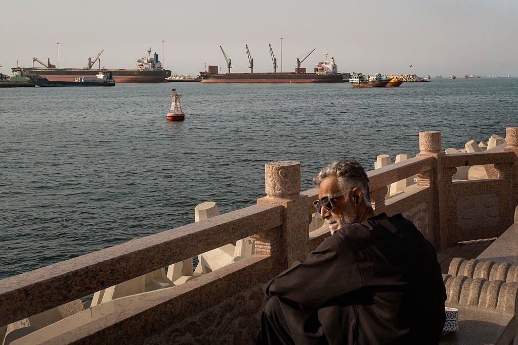 A man with grey hair, black clothing and sunglasses looks on from a stone observation point in Oman at the Strait of Hormuz. In the distance are at least five boats and a buoy floating on the water.