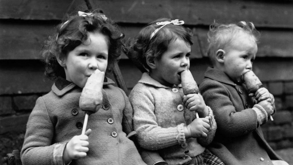 This black-and-white photograph shows three young children sitting side by side outdoors in front of a wooden plank wall. Each child is holding and eating a root vegetable on a stick. The children are dressed in coats or cardigans suitable for cool weather, and two of them wear ribbon bows in their hair.