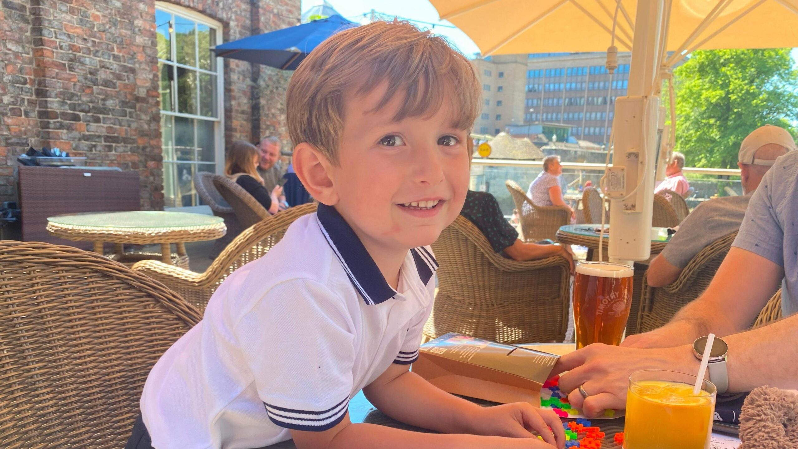 Young boy in white polo t-shirt smiles into camera as he leans over the table with an orange juice in front of him.