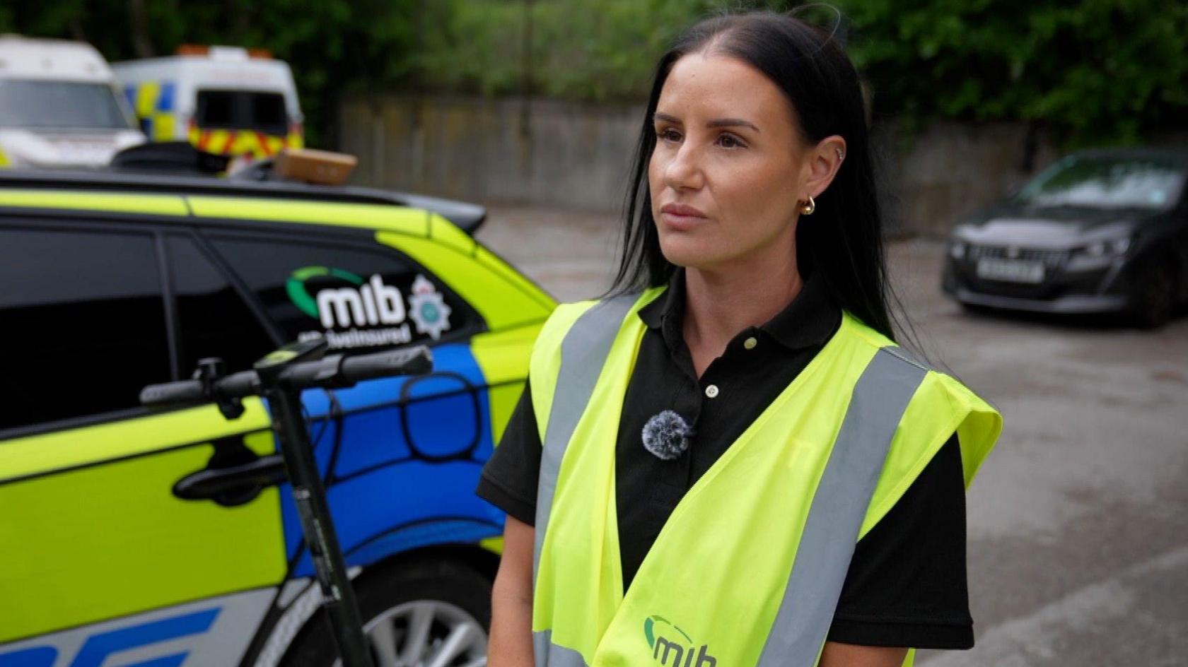 Hayley Sutcliffe  from the Motor Insurers' Bureau is standing in front of a police car and an e-scooter, she has dark shoulder length hair and is wearing a high-vis vest.