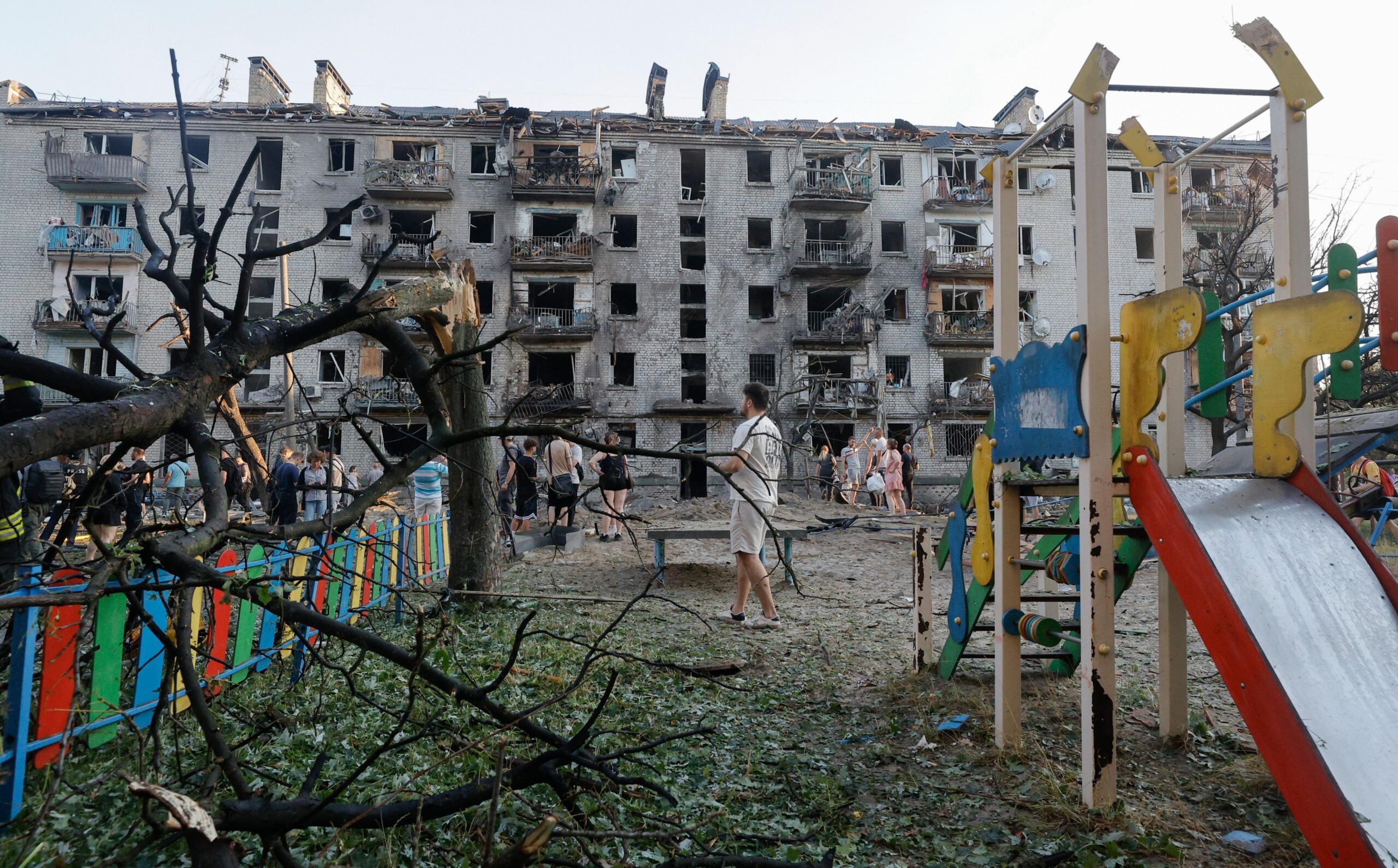A partially-damaged playground slide in yellow, red and green stands on the right of the frame. A tree split at the trunk is bent over a fence also painted in red, green, yellow and blue. In the background tens of residents walk around the while a partially-destroyed building sits in the background.
