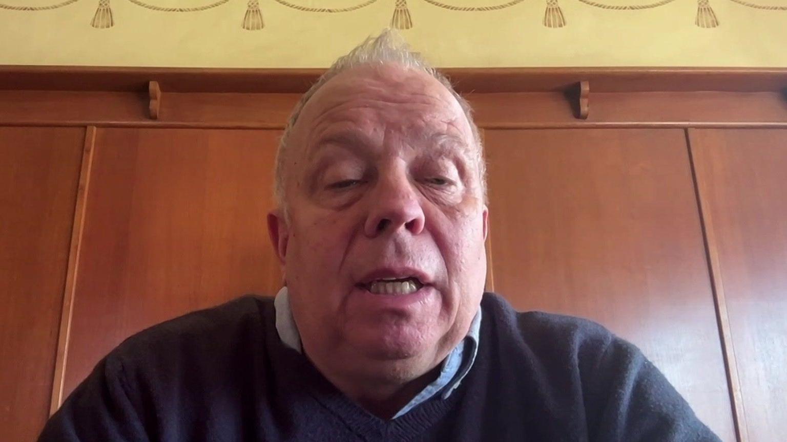 John Stiles is seen on a video link, talking to a reporter. He is middle-aged, has short light-coloured hair, and is wearing a shirt and jumper