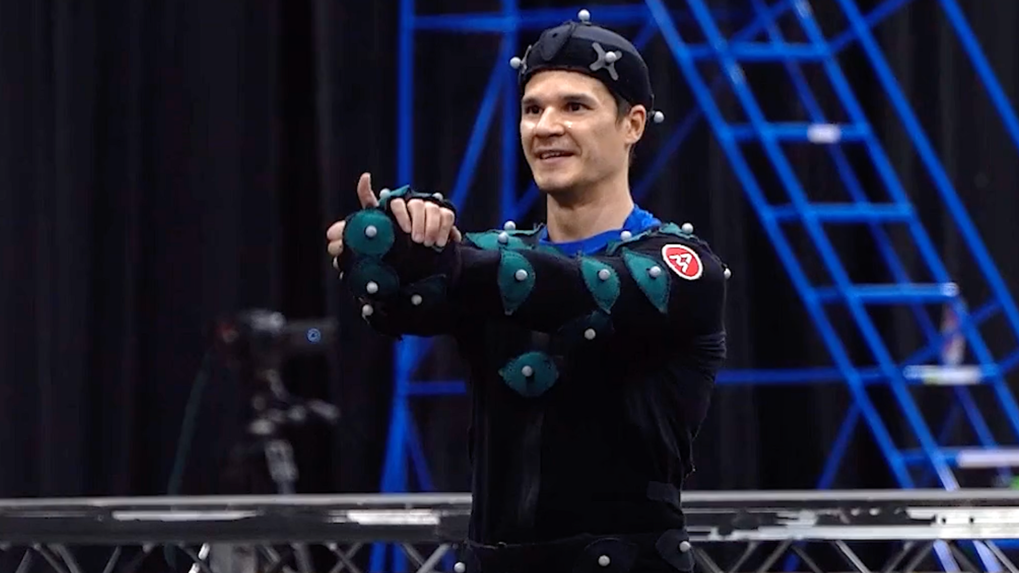 A man stood with his thumb up looking past the camera. He is wearing black overalls with green patches on where white balls sit. He is also wearing a hat with white dots on.