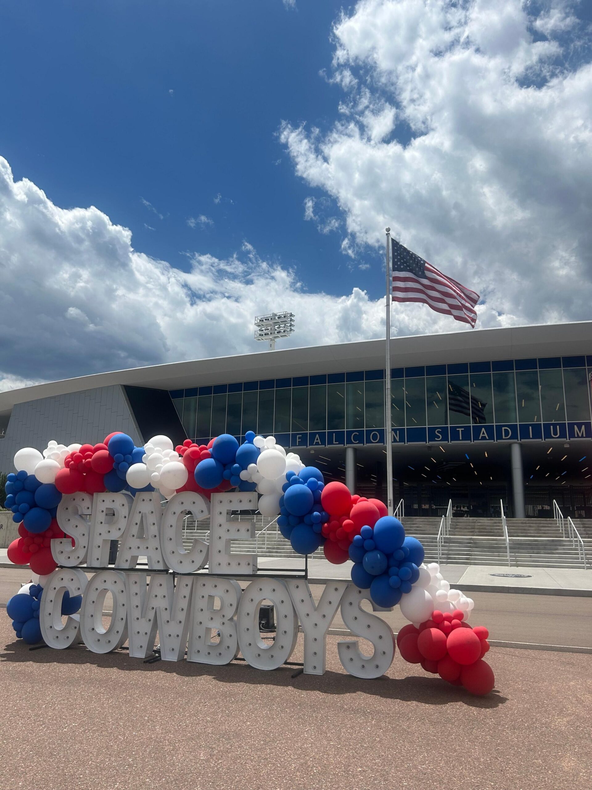 A huge white shine reading "Space Cowboys" in block capital letters stands covered in an arch of red, white and blue balloons outside Falcon Stadium, whose name and lights can be seen in the background as an American flag waves on a pole between the Space Cowboys sign and the steps up to the stadium doors