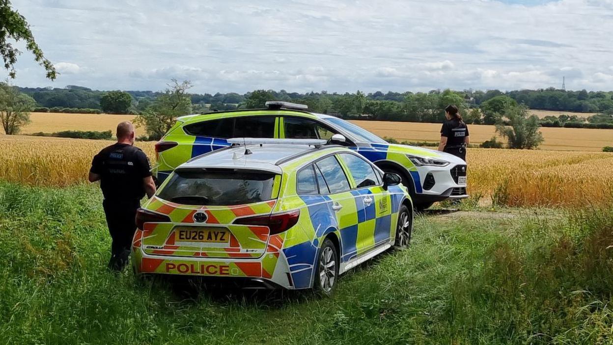 Two police cars parked by the side of a large field. Two officers are looking into the field beyond them.