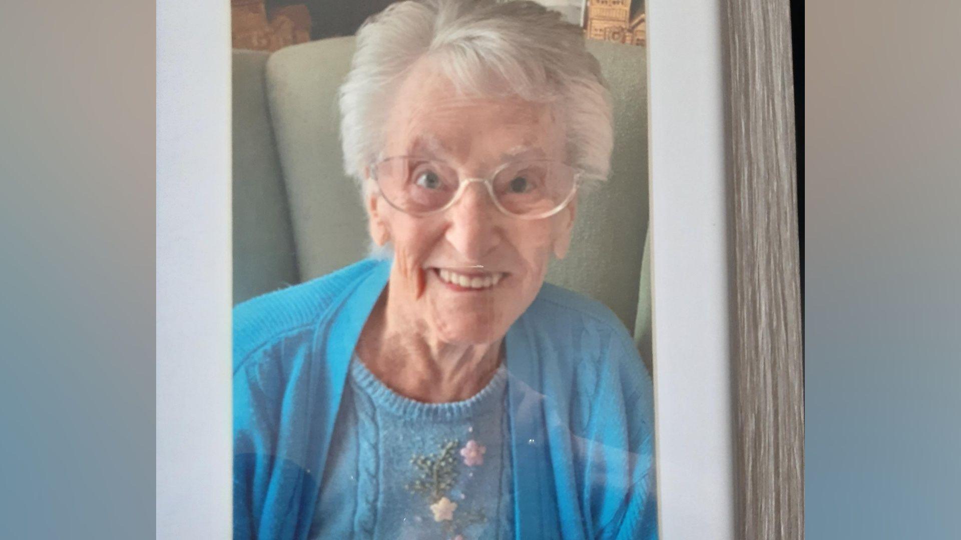 Joyce Moulton pictured sitting on a chair smiling. She has grey hair and is wearing a blue cardigan and glasses.