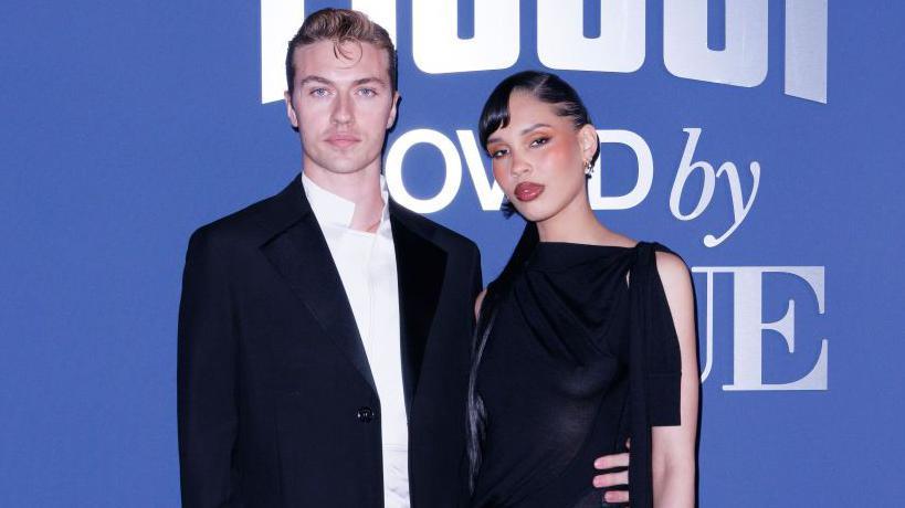 Married couple Lucky Blue Smith and Nara Smith stand against a blue backdrop in formal wear. Nara is wearing a black dress with strands on each side and Lucky wers a white shirt and black blazer.