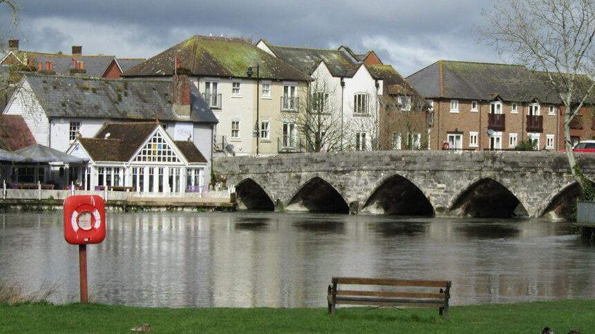 An old stone bridge with arches spans the River Avon at Fordingbridge. A bench and life ring are on the grass-covered near bank. The far bank is clustered with houses.