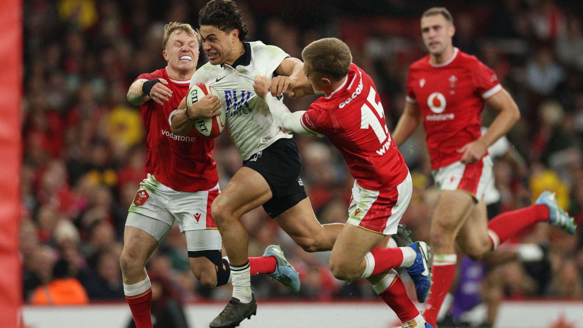Fiji's Caleb Muntz in black and white kit bursts through the tackles of Wales players in red and white