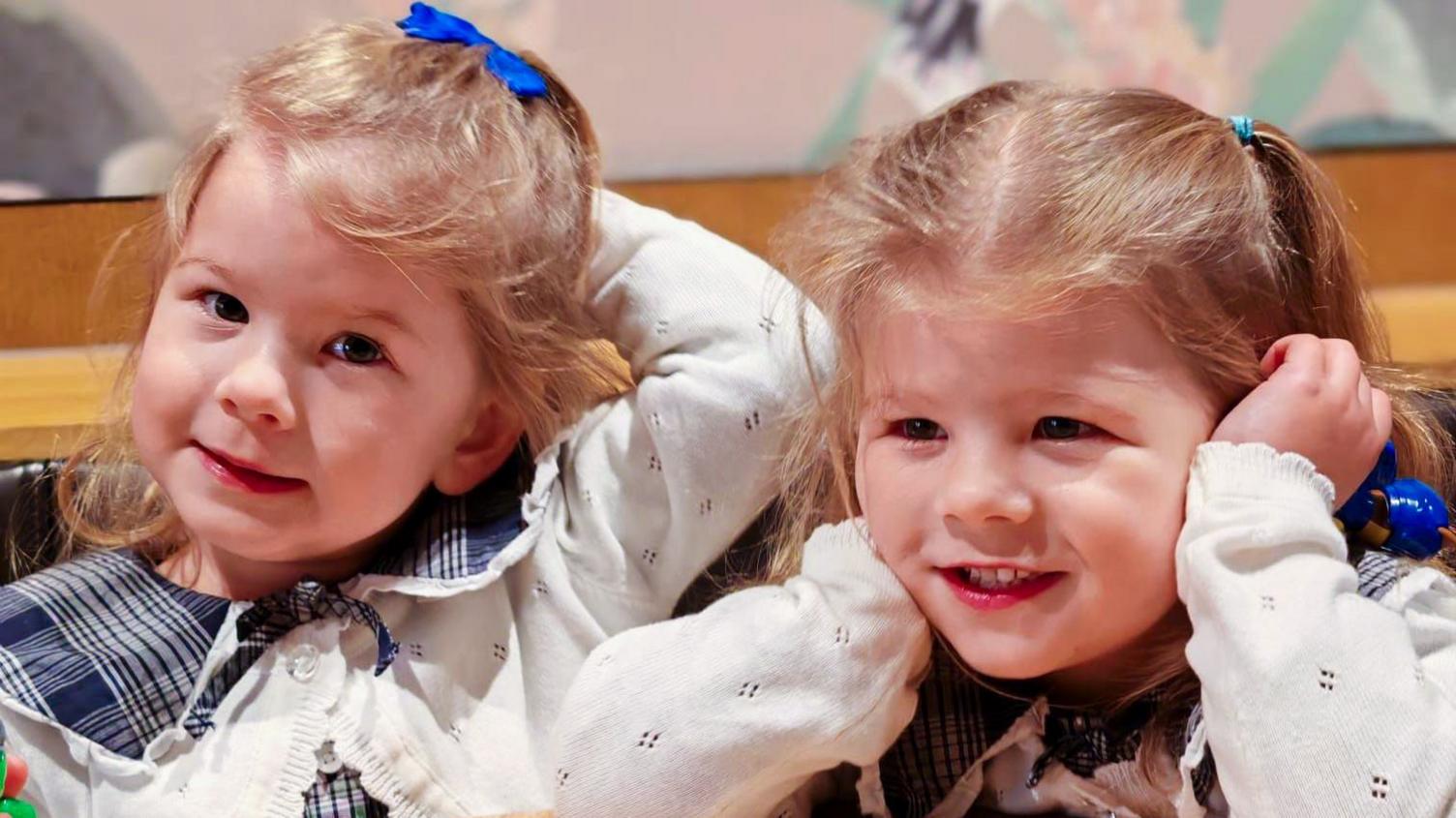 Nancy and Margo are photographed close up - their faces and blond hair in bunches are visible, and they are smiling. They are wearing the same clothes - a blue blouse and white cardigan.