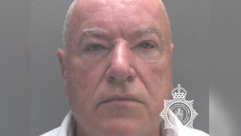 A police mugshot of Neil Foden. He has short white hair and is looking directly into the camera. He is almost entirely bald, with fine, grey very cropped hair on the sides. The background is all grey and he is wearing a white collared shirt