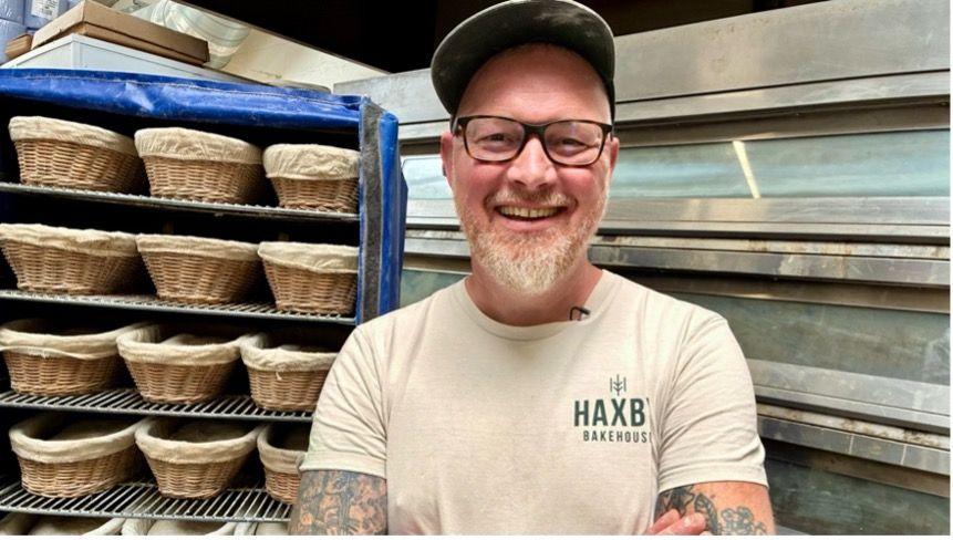 Phil Clayton, a man wearing a hat and glasses and with tattoos on his arms, is smiling broadly as he stands in front of an industrial over. To the left of the image are racks full of baskets.