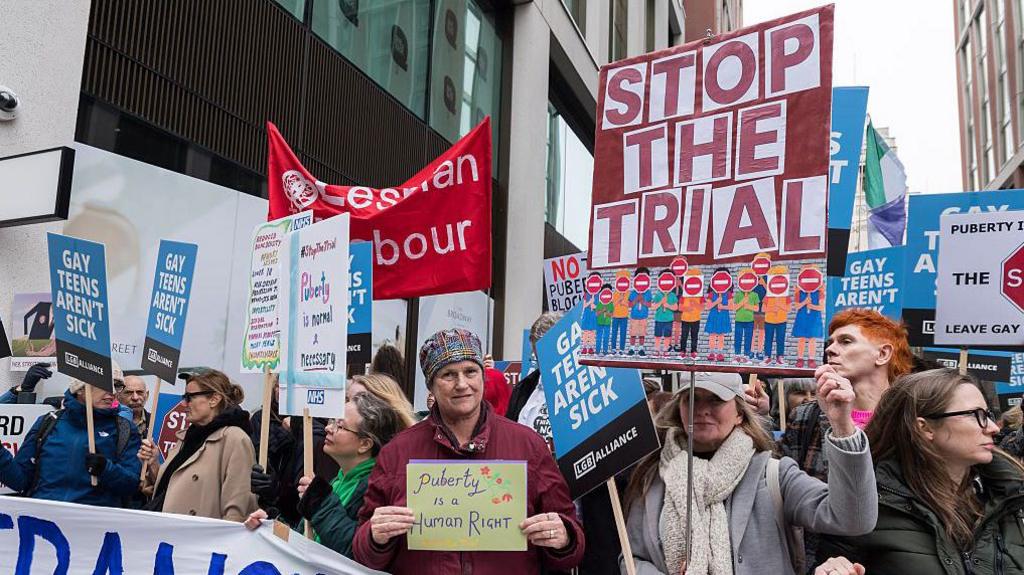 Demonstrators pictured in central London in December 2025 protesting against the clinical trial. One sign reads "Stop the trial" and another reads "puberty is a human right".