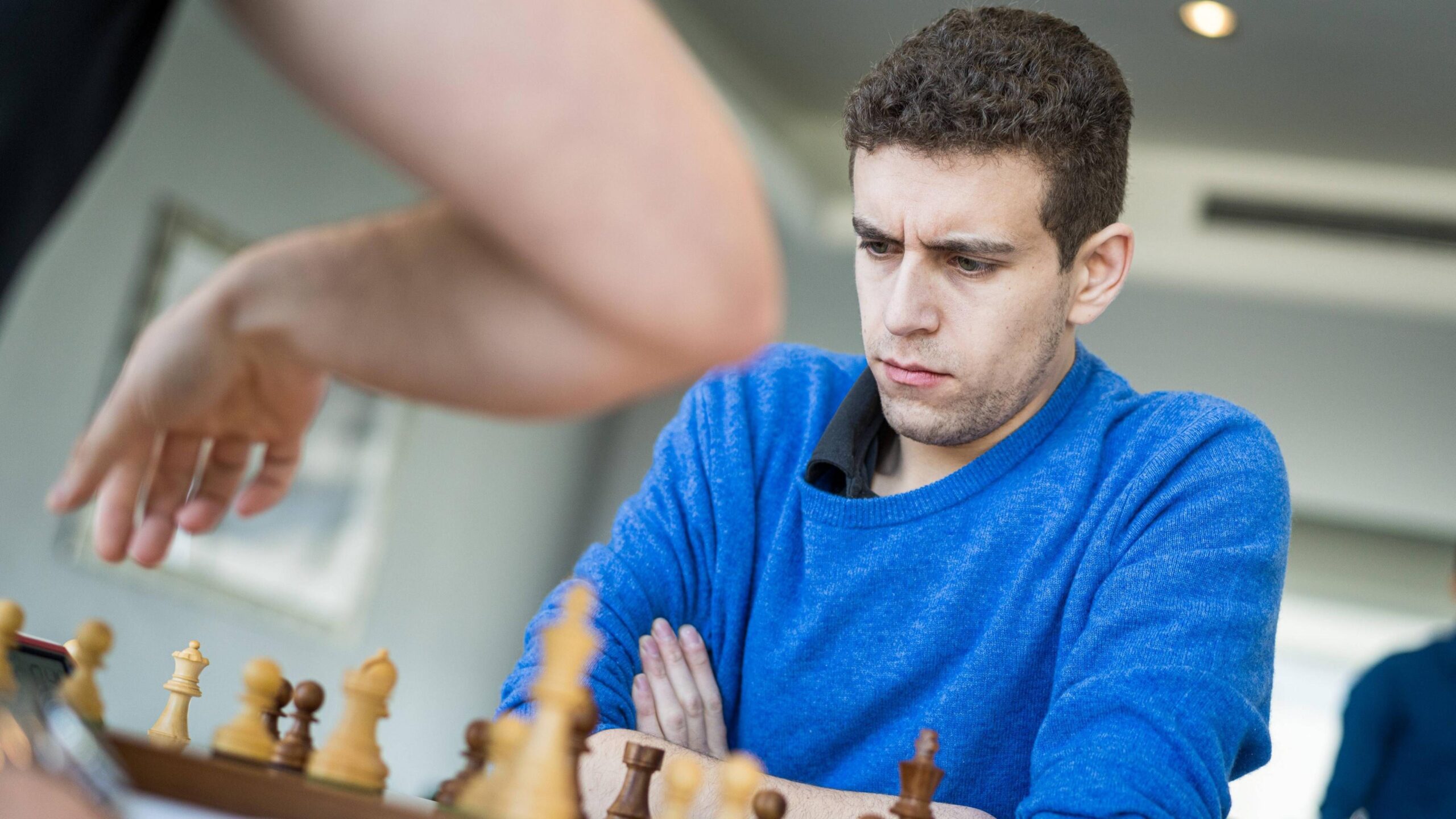 Naroditsky sits in front of a chess board with his arms crossed. He is wearing a navy shirt and a blue jumper.