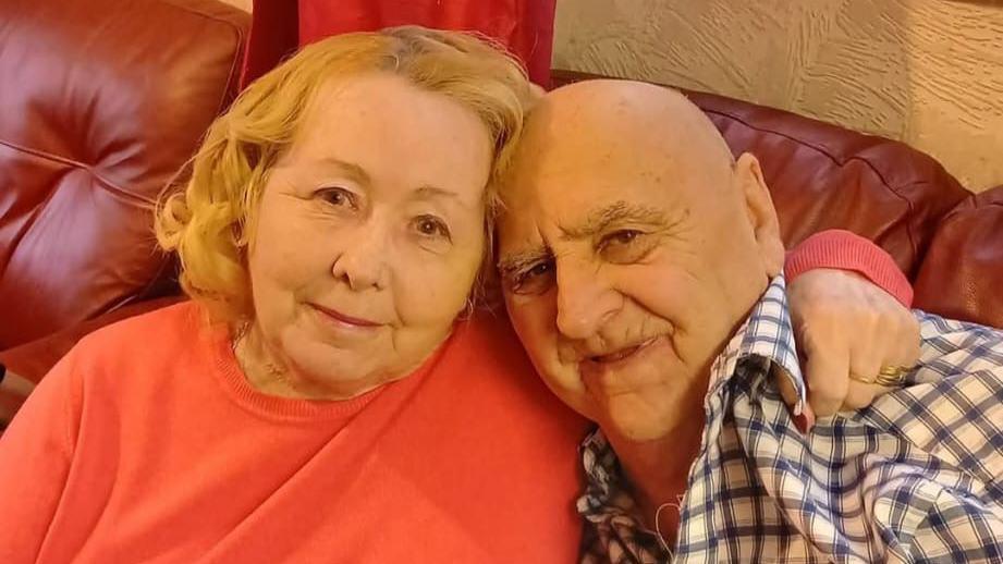Elizabeth and her husband John are photographed sat on a red sofa from the shoulders up. Elizabeth has her arm around his shoulder.