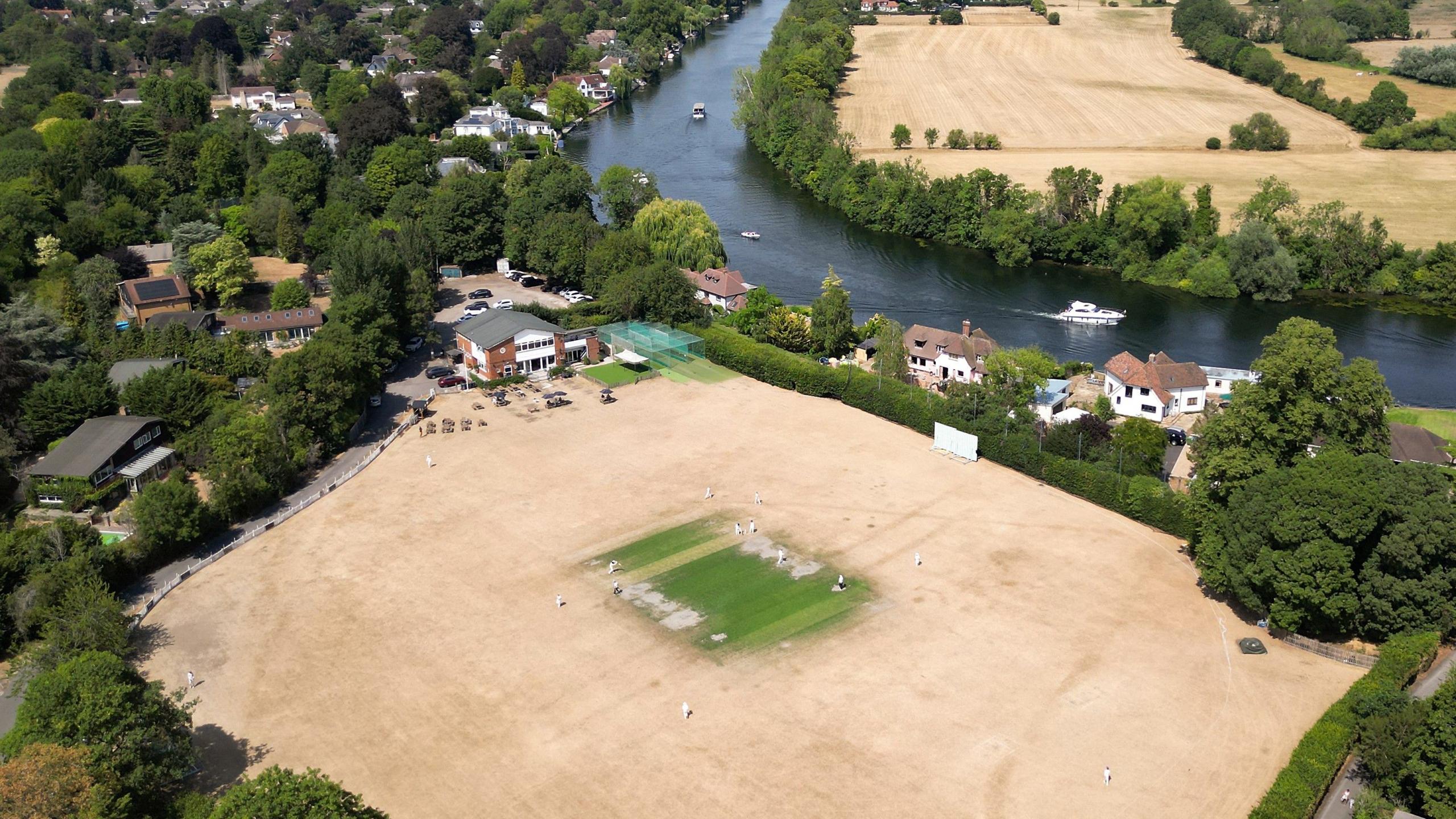 A match taking place on a cricket field parched pale brown by drought, except for a green centre square. A river flows past