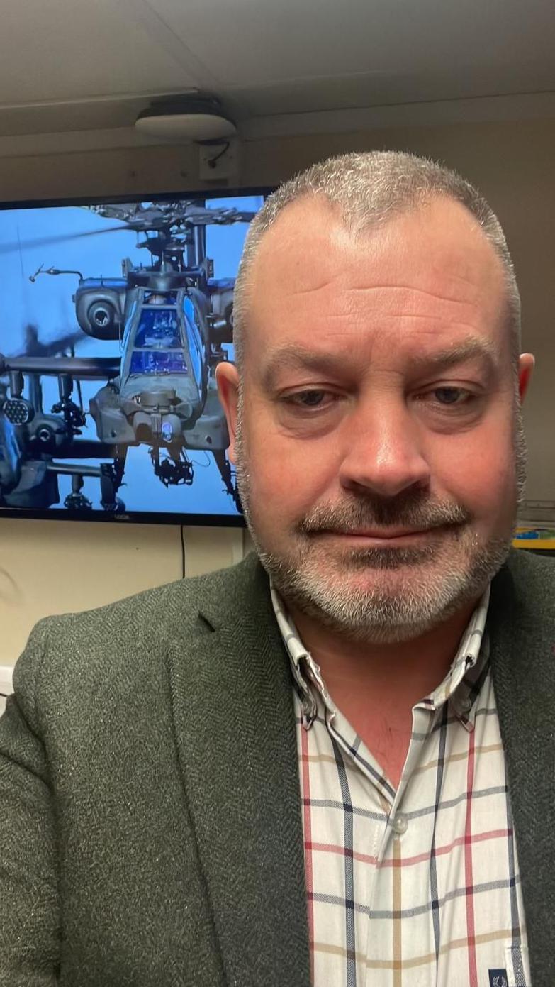 Griffiths dressed in a checked short and blazer standing in front of TV screen with a helicopter on it